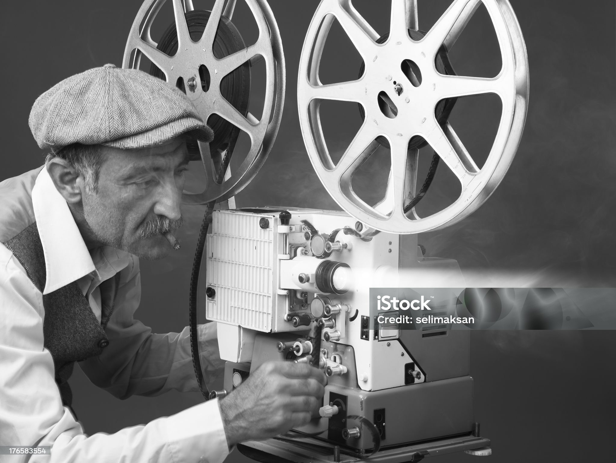 Senior man wearing shirt,waist and flat cap starting old fashioned film projector.He is placed on the left side of the frame.Smoke ad light bean come out of projector lens.The photo is black and white and has horizontal composition.Shot with a Full frame DSLR camera.